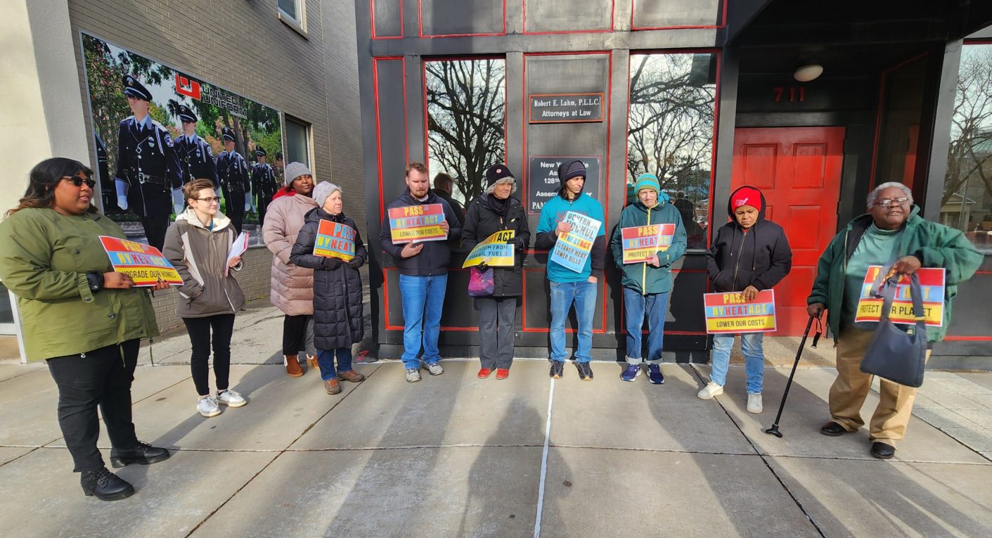 Activists stand in a semi-circle facing the camera listening to the press conference speaker, holding signs saying "Pass the NY HEAT Act. Upgrade our buildings" and "Governor Hochul. NY HEAT Act. Cleaner Heat Lower Bills".